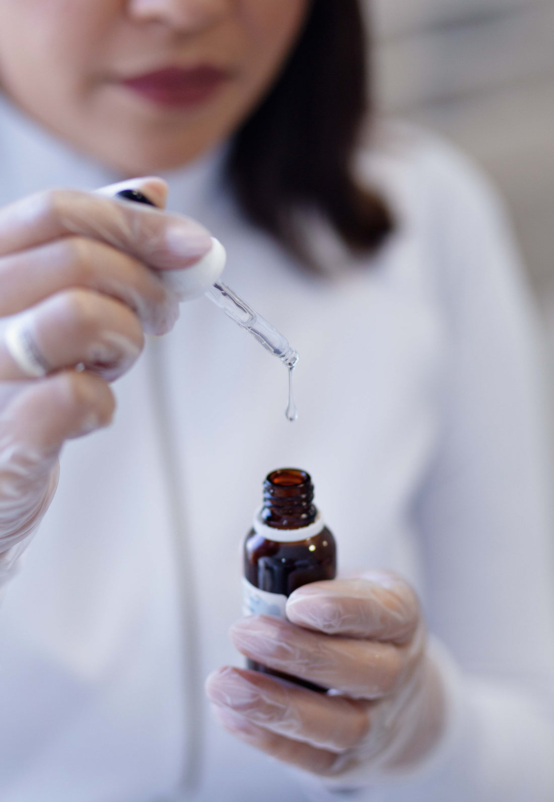 Person in gloves using dropper with clear liquid serum from amber bottle.
