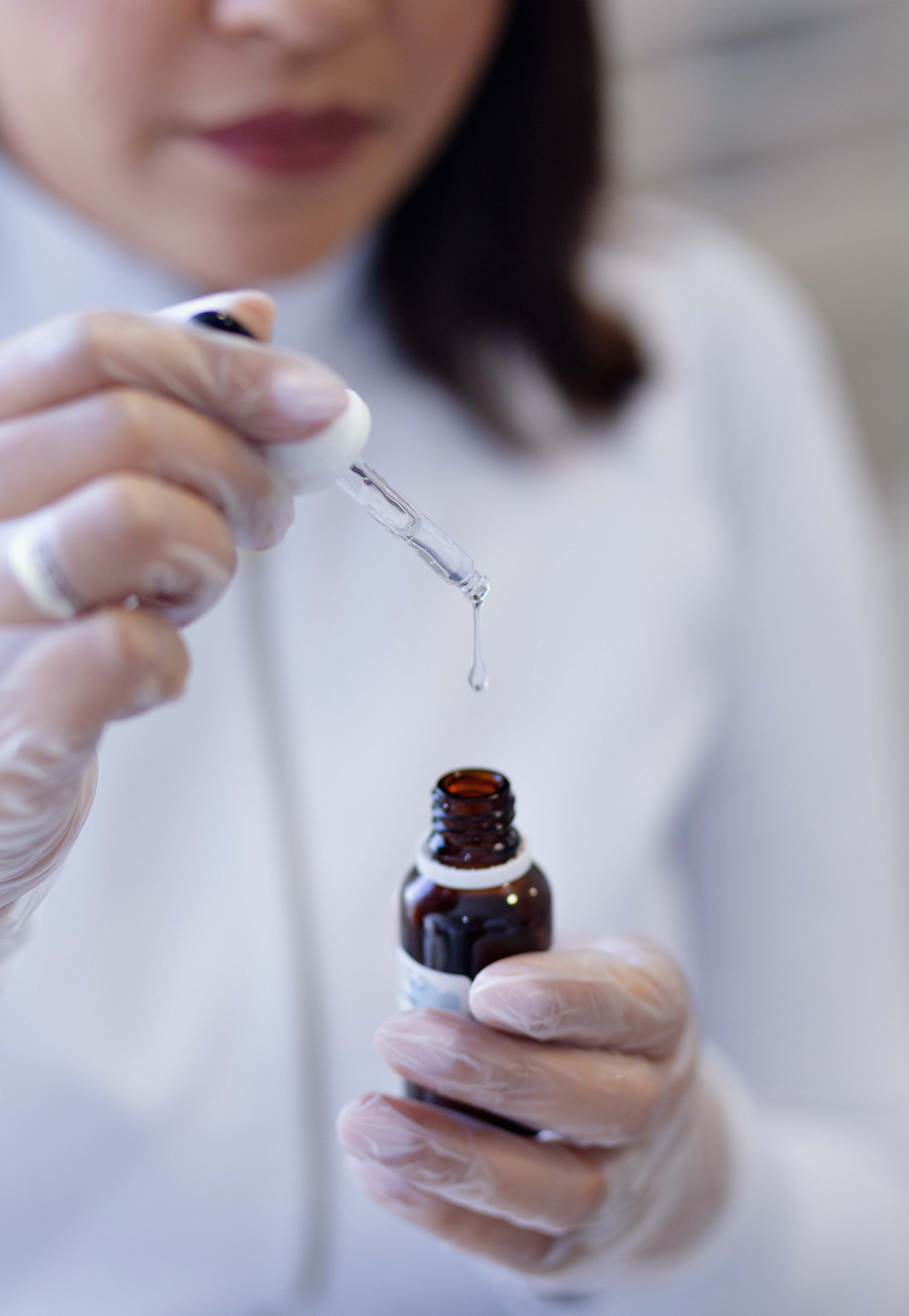 Person in gloves using dropper with clear liquid serum from amber bottle.